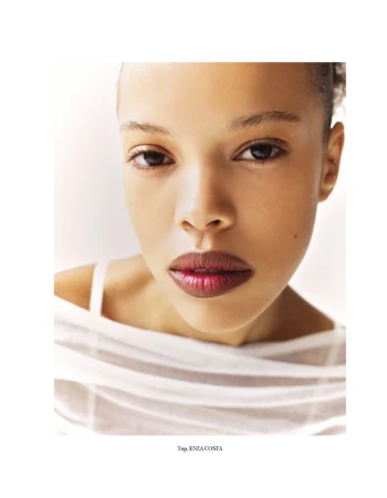 Close-up portrait of a young adult woman with fair skin, wearing a sheer white top against a plain white background.