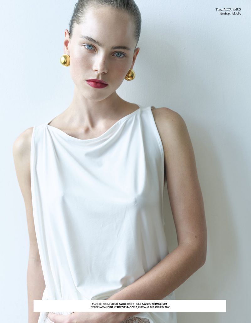 Portrait of a fair-skinned woman with red lipstick, wearing gold earrings and a white top, gazing directly at the camera against a white wall.