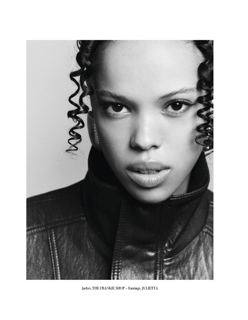 Close-up portrait of a young adult woman with curly hair wearing a dark leather jacket against a neutral background in black and white.