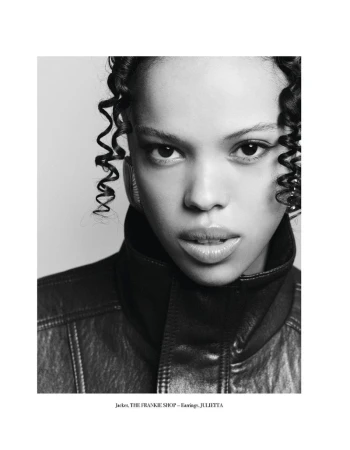 Close-up portrait of a young adult woman with curly hair wearing a dark leather jacket against a neutral background in black and white.