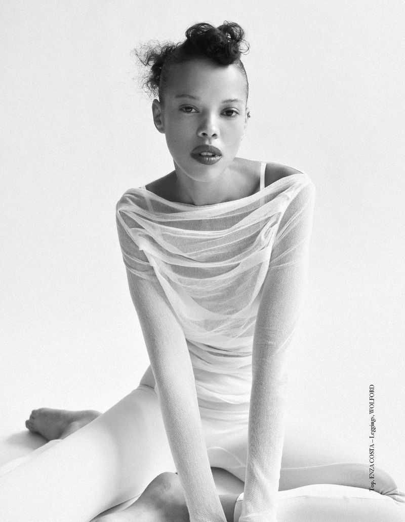 Black and white studio shot of a Black female model with sheer draped top and leggings sitting on the ground looking at the camera.