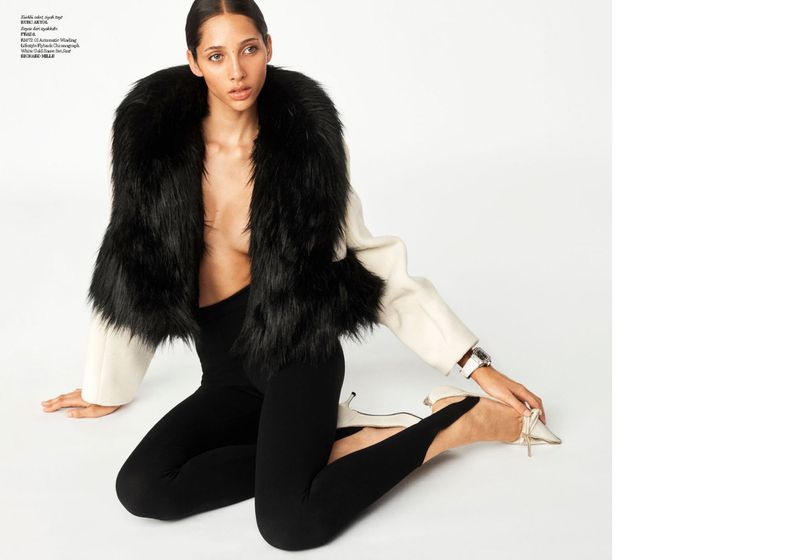 Fashion shot of woman in black leggings and jacket with fur collar sitting on a white floor, posing with hands and feet.