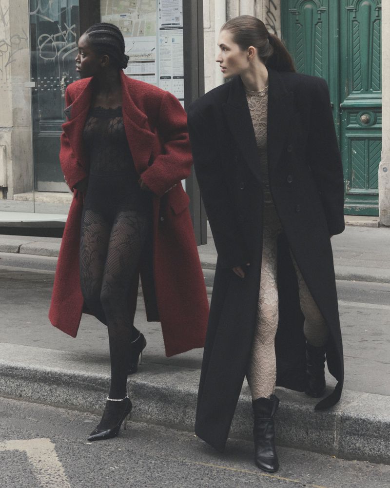 Two fashion models on city street wearing coats over lace bodysuits and hosiery, one in red coat and heels, the other in black coat and boots.