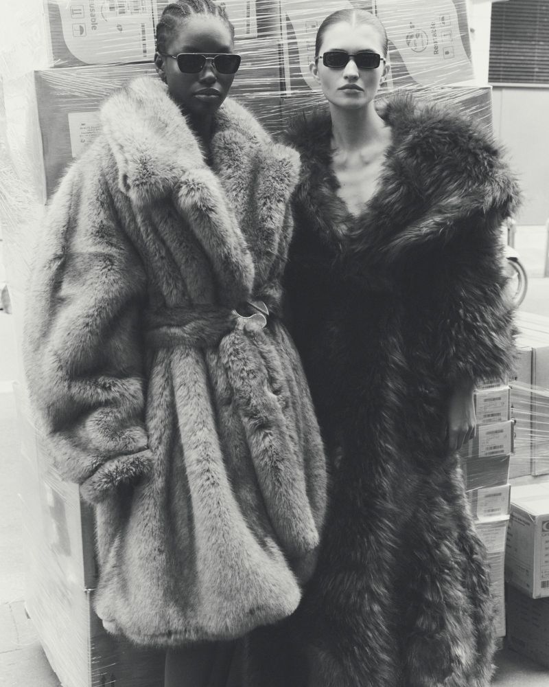 Two women wearing sunglasses and fur coats pose in front of stacked boxes in a black and white fashion shot with a shallow depth of field.