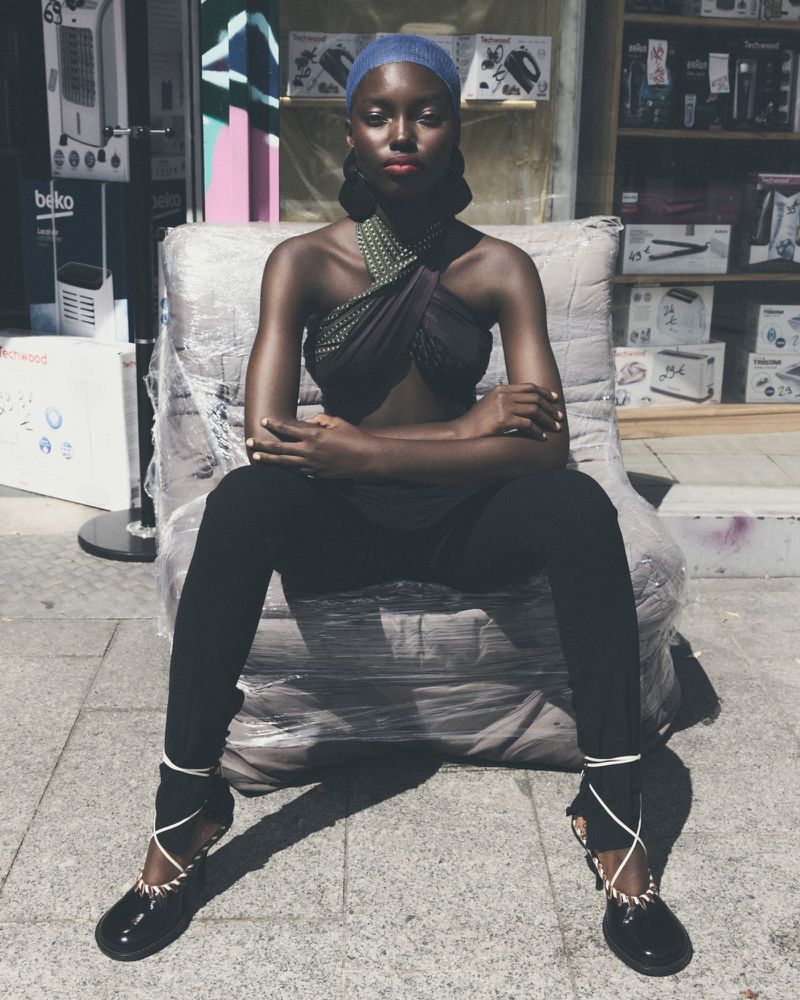 Elegant Black woman sitting outdoors in black leggings, top, and ballet flats with wraparound ties, posing in front of a storefront.
