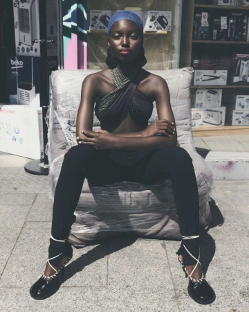 Elegant Black woman sitting outdoors in black leggings, top, and ballet flats with wraparound ties, posing in front of a storefront.