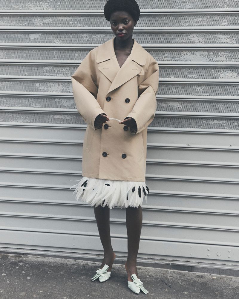 A black female in a tan double-breasted coat and feathered skirt poses against a gray corrugated metal background, showcasing a fashion-forward look.