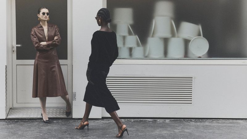 Two fashion models showcase chic midi dresses in an urban setting; one in brown leather, the other in black velvet, with minimalistic heels.