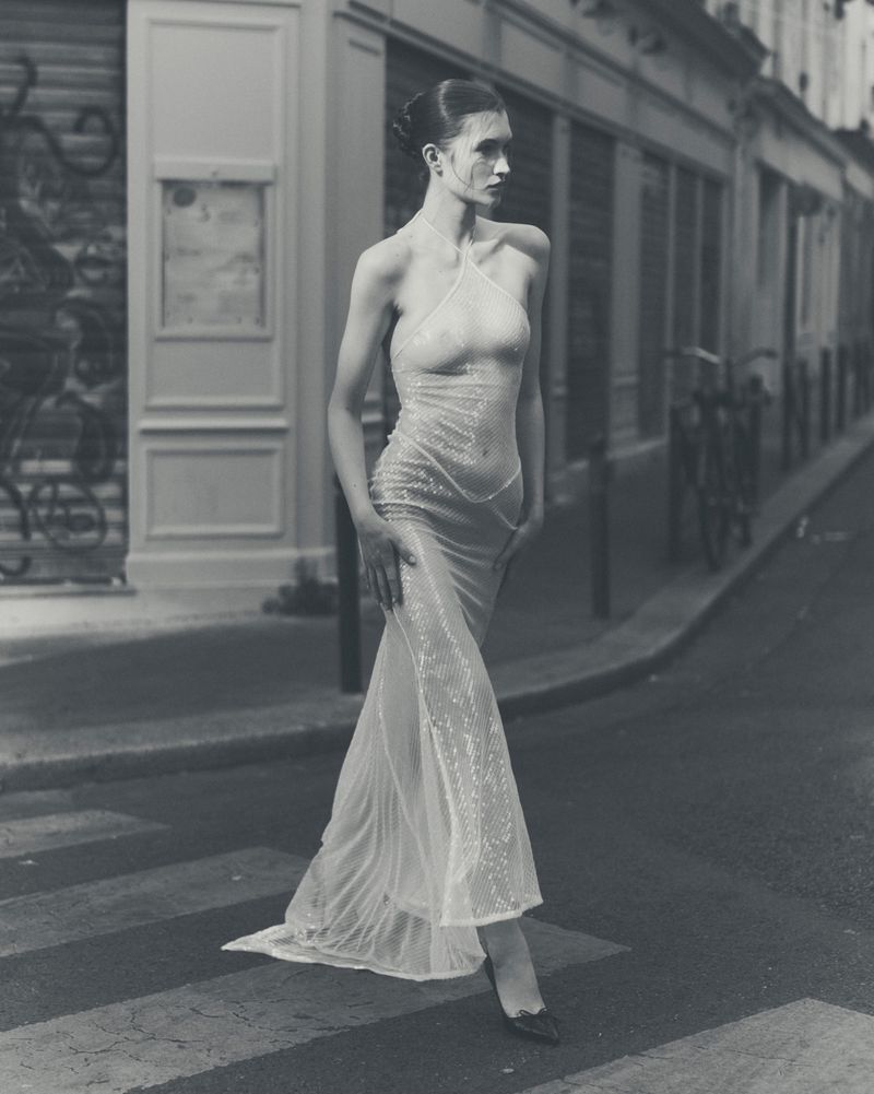 A slender woman in a sheer gown walks across a zebra-striped crosswalk in an urban setting, with European architecture in the backdrop, in monochrome.