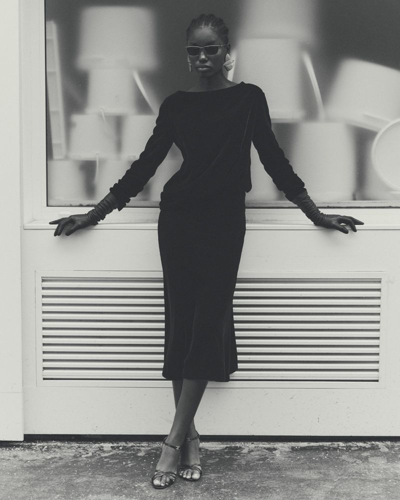 Monochrome fashion shot of an adult woman in a long-sleeved black dress, gloves and sandals, leaning against a light wall and background.