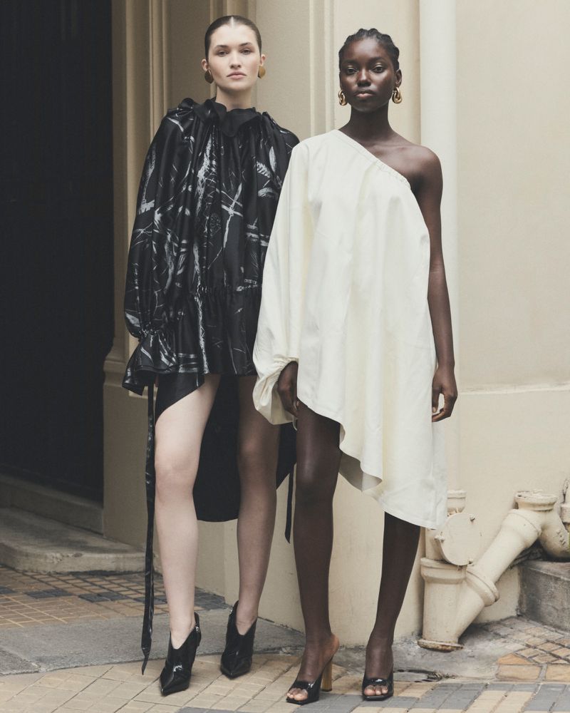 Two women stand in stylish black and white dresses, accentuated by statement earrings and sleek heels, in front of a neutral wall.