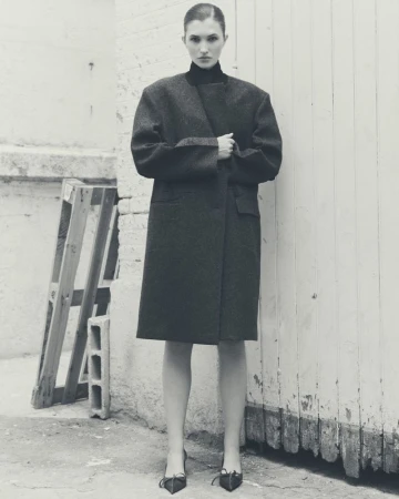 Monochrome full-length shot of a female fashion model in a dark overcoat and heels posing in front of a weathered white brick wall.
