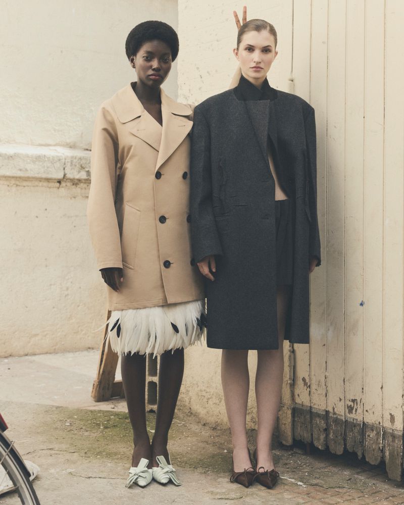 Two women pose in outerwear against an urban background. The woman on the left wears a beige coat with feather details.