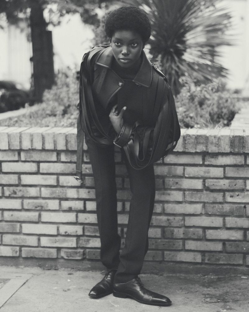 An adult female with a serious expression is photographed in a black and white outdoor portrait wearing fashionable outerwear and dress shoes.