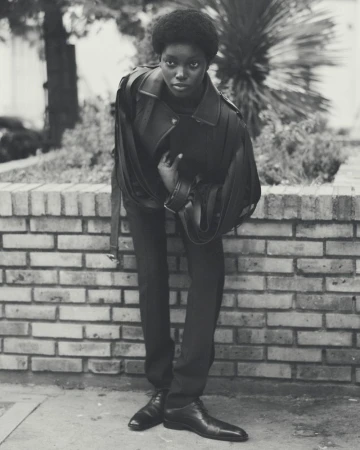 An adult female with a serious expression is photographed in a black and white outdoor portrait wearing fashionable outerwear and dress shoes.