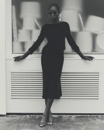 Miriam Ntuyang wearing a black long-sleeved midi dress, opera gloves, and sunglasses leaning against a white architectural facade.
