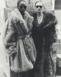 Two female models wearing oversized faux fur coats and dark sunglasses posing in front of stacked plastic-wrapped boxes.
