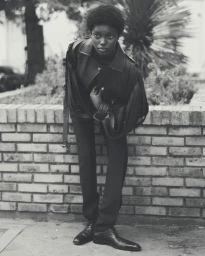 Monochrome portrait of Miriam Ntuyang leaning forward in a dark jacket, tailored trousers, and dress shoes against a brick planter.