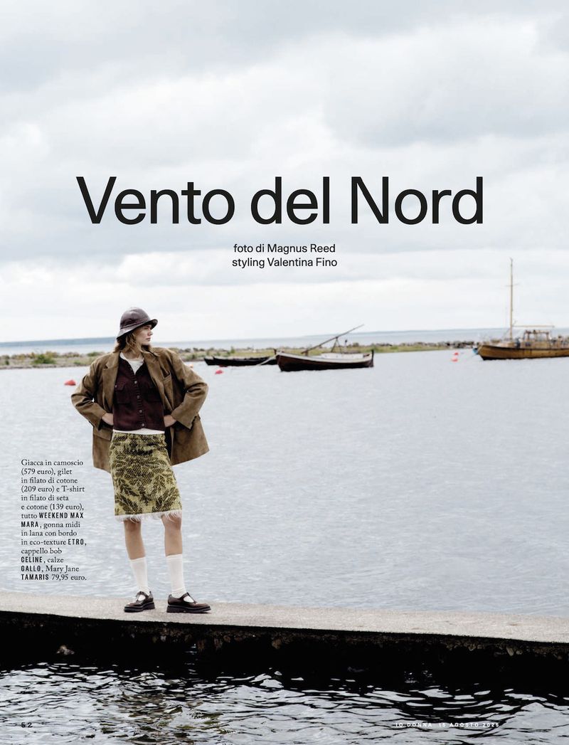 Fashion editorial shot of a woman in a brown jacket and patterned skirt stands on a pier with boats and cloudy sky in the background.