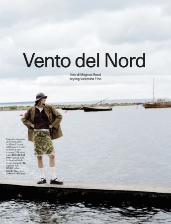 Fashion editorial shot of a woman in a brown jacket and patterned skirt stands on a pier with boats and cloudy sky in the background.