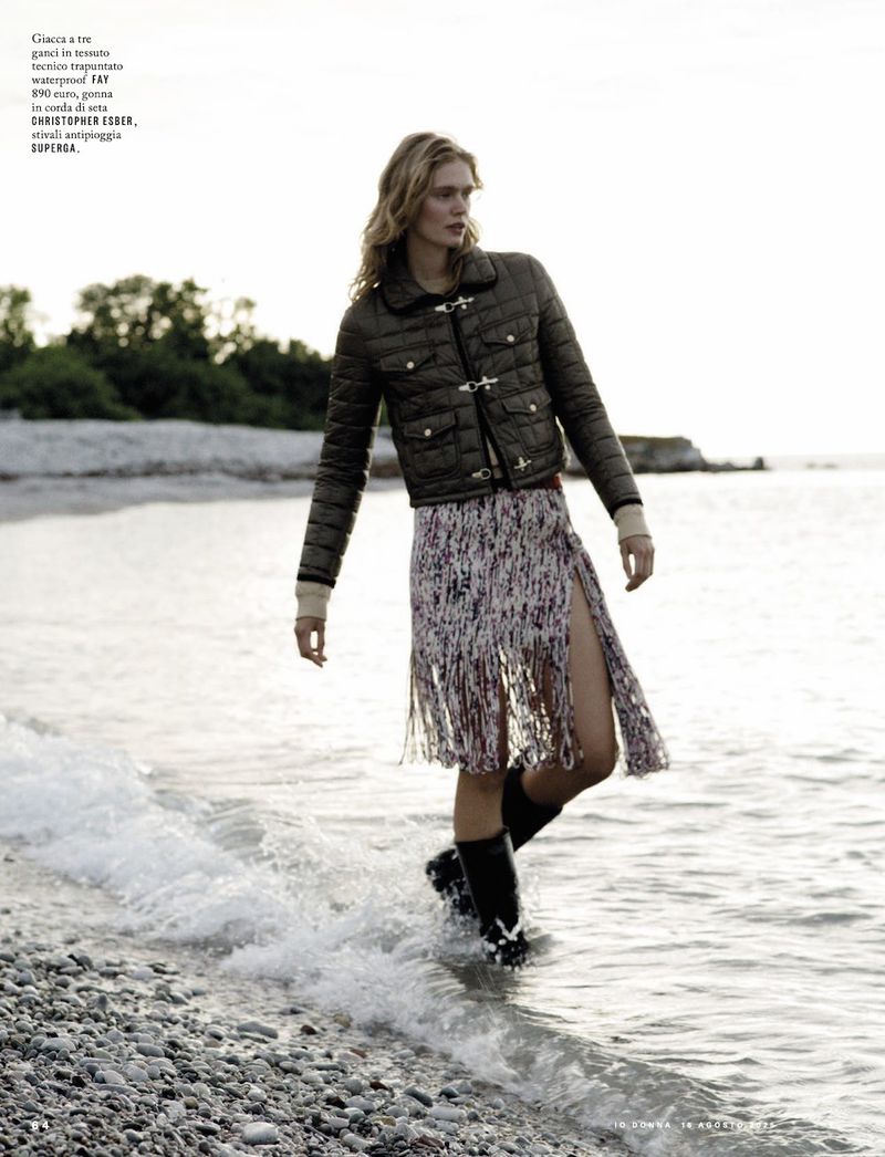 A blond woman in a puffer jacket, patterned skirt, and rainboots walks on a rocky beach as waves wash around her feet.