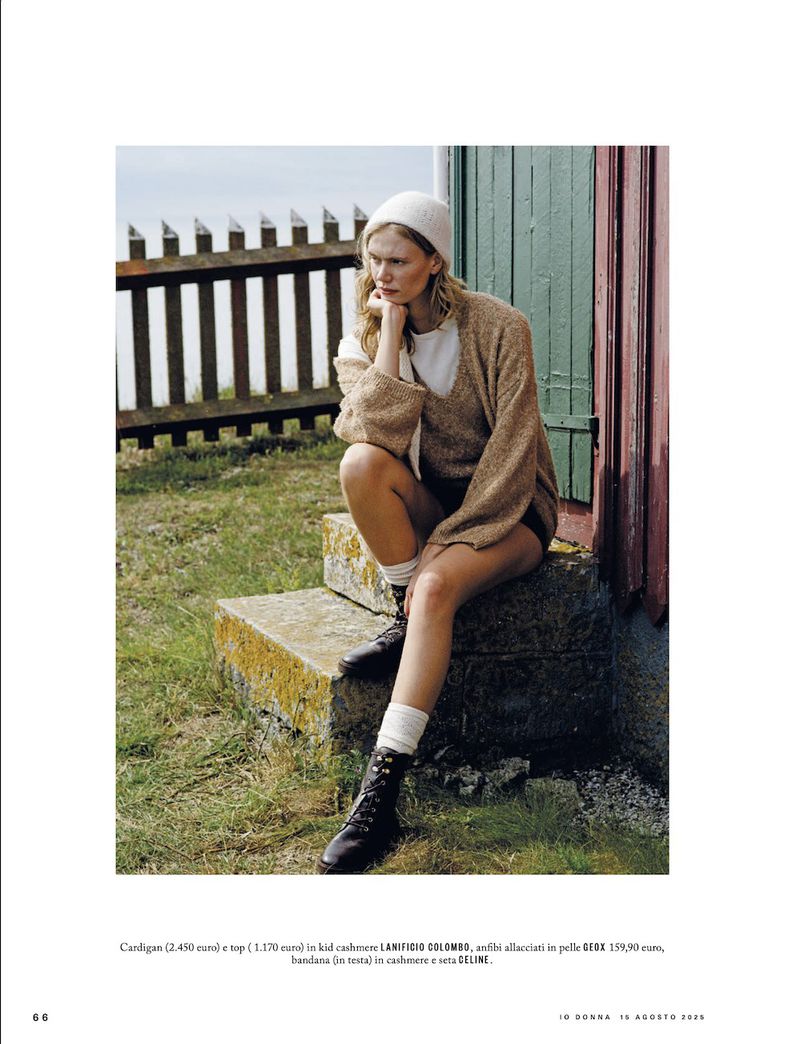 A woman in a cardigan and beanie poses outdoors on stone steps next to a building with a weathered, painted wooden fence in the background.