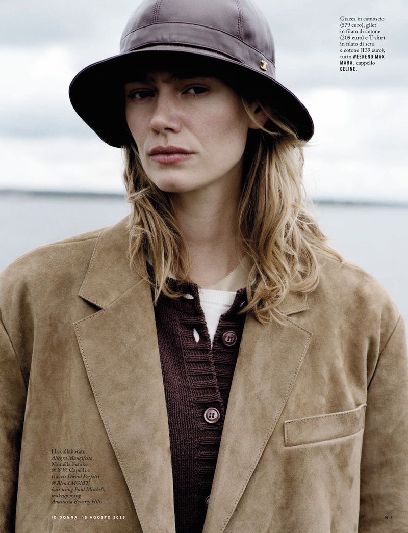 A woman with long blonde hair wears a brown hat and tan coat, standing outdoors with a calm expression against a blurred seascape backdrop.
