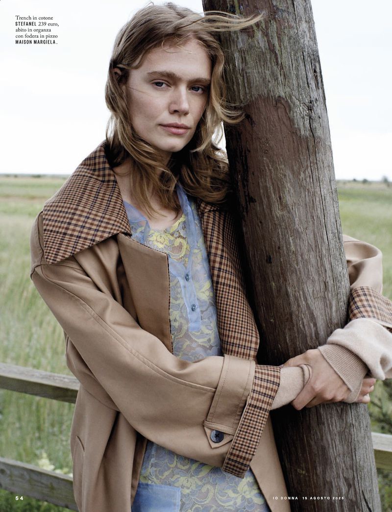 Woman in a tan trench coat poses outdoors near a weathered wood pole, with windblown hair and a serene expression, in soft, natural light.