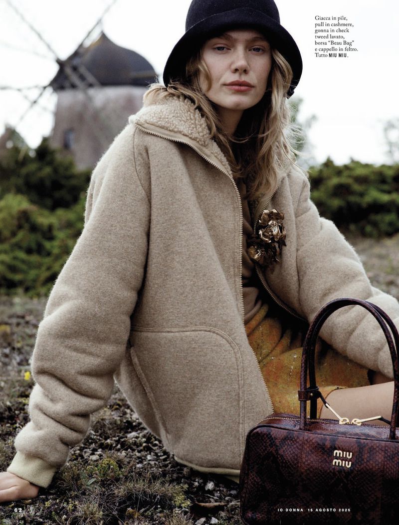 A woman wearing a hat and jacket is seated outdoors with a purse, a windmill in the background, fashion photography, lifestyle editorial.