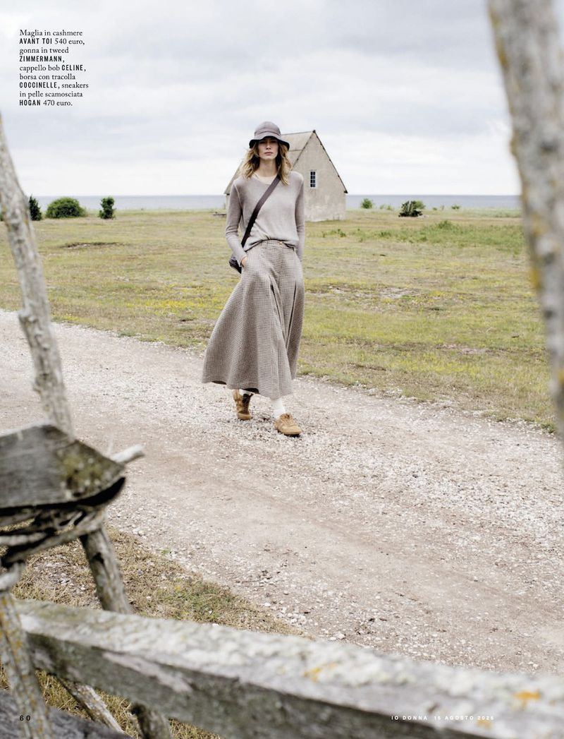 Woman in long beige skirt and sweater walking on path near beach and house under overcast sky, framed by weathered wooden fence.