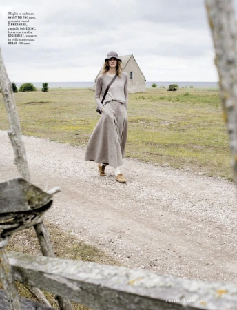 Woman in long beige skirt and sweater walking on path near beach and house under overcast sky, framed by weathered wooden fence.