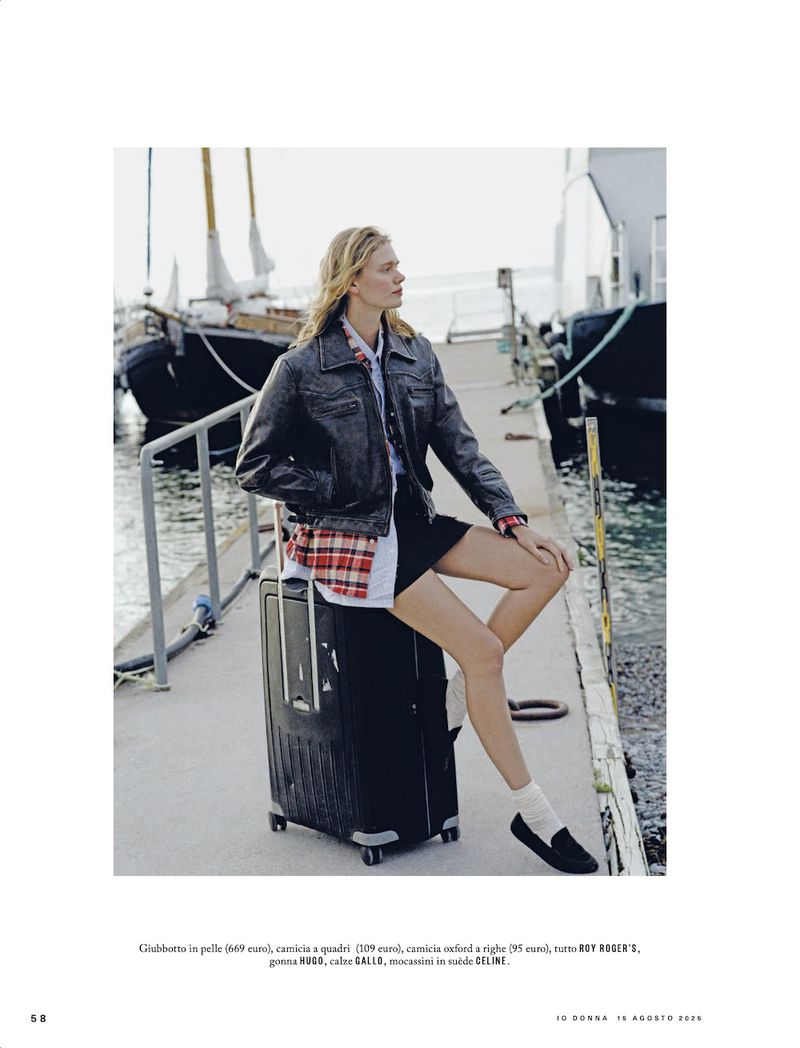 Fashion shot of a woman in a black jacket and skirt sitting on a suitcase near the sea, with a sailboat in the background.