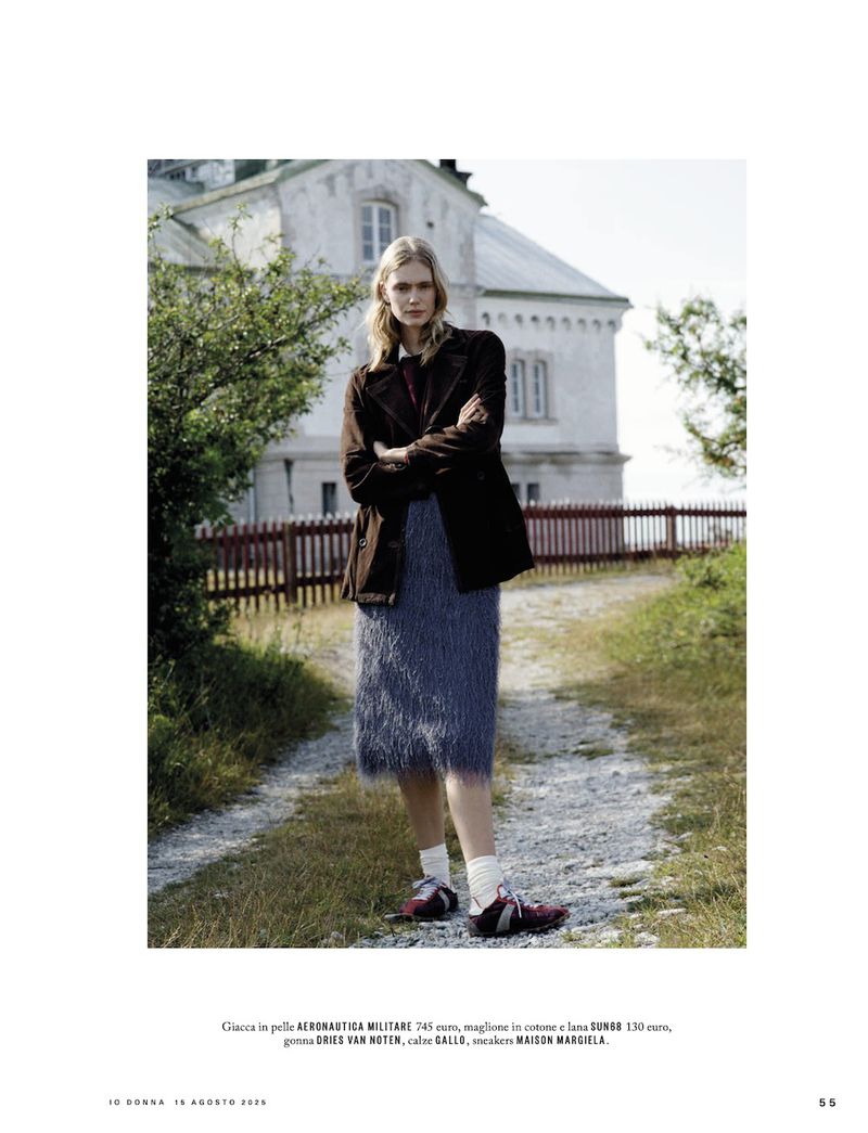 Blonde woman in a brown jacket and blue skirt stands outdoors in front of a rural building on a sunny day for a fashion editorial.