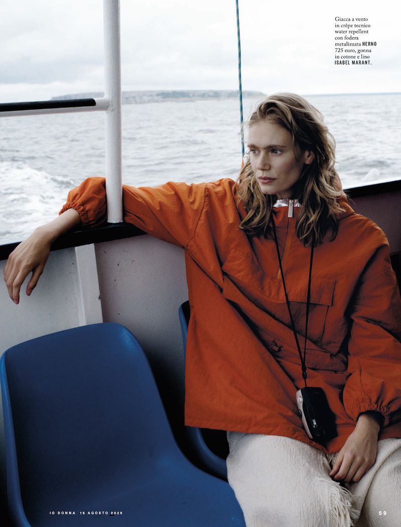 A blonde woman in an orange jacket and white pants sits on a blue chair on a boat, looking out at the ocean on a cloudy day.