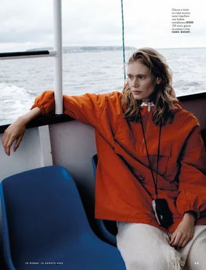 Model Femke Bloem in a bright orange windbreaker seated on a boat deck with blue plastic chairs against an open ocean background.