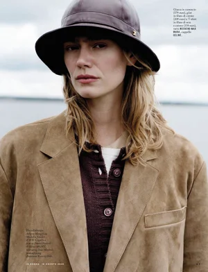 Model Femke Bloem wearing a brown suede blazer, dark knit cardigan, and a brown leather hat against an overcast sky and water.