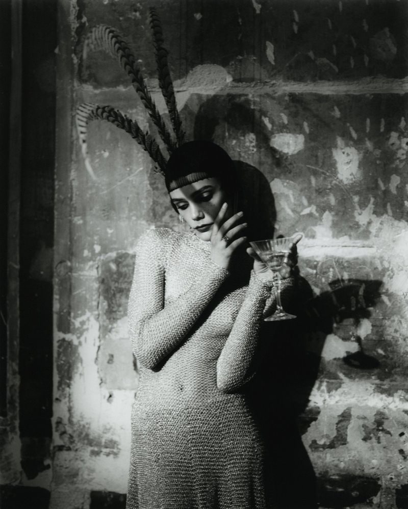 Monochrome portrait of a slender woman in a feathered cap and chainmail dress holding a cocktail glass against a textured wall.