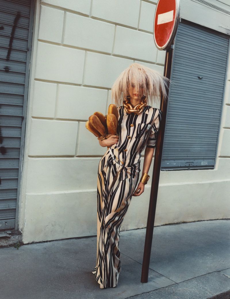 Fashion model in a striped maxi dress and blonde wig posing next to a stop sign on a city street, accessorized with a gold necklace.