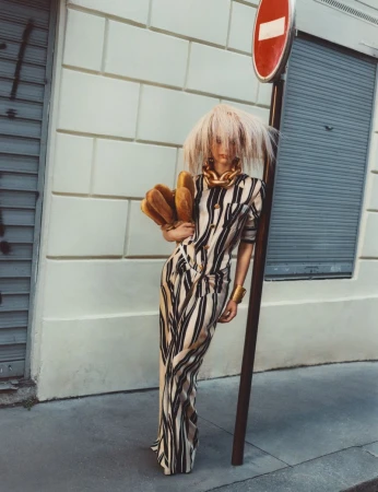 Fashion model in a striped maxi dress and blonde wig posing next to a stop sign on a city street, accessorized with a gold necklace.
