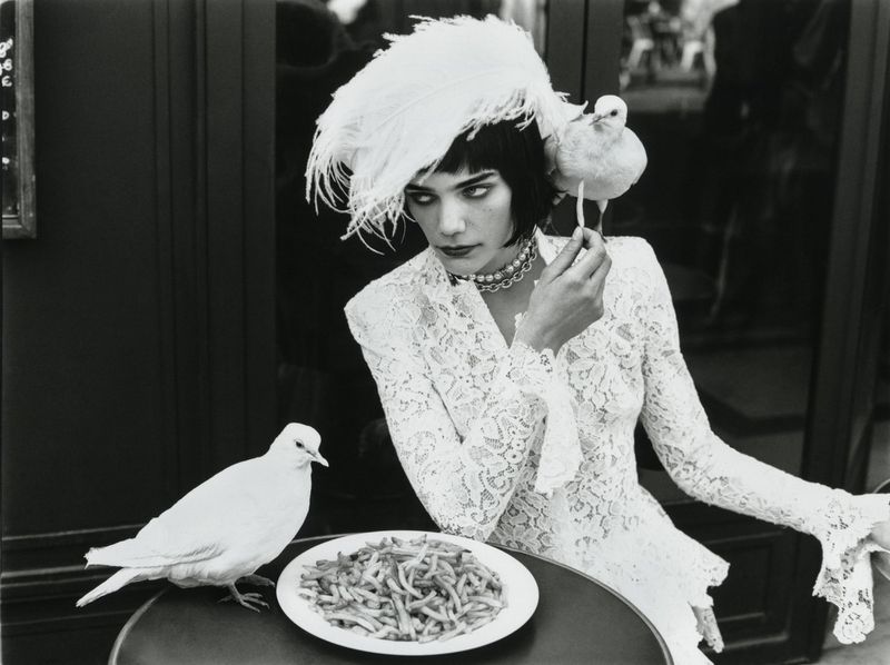 Monochrome portrait of a woman in a lace dress, adorned with feathers and birds, seated at an outdoor café table with fries.