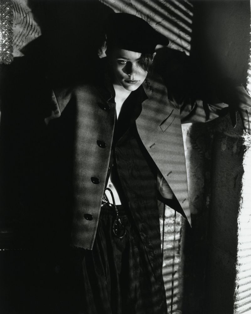 Monochrome portrait of a person wearing a beret and jacket in a room with light filtering through window blinds creating shadows.