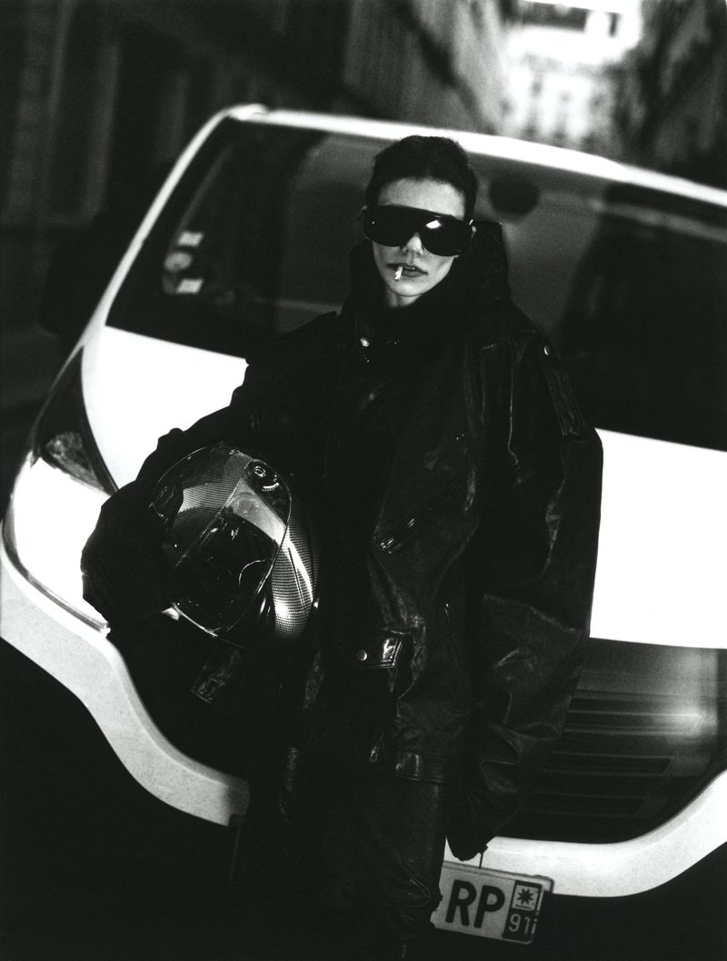 Monochrome shot of a stylish adult leaning on a car, cigarette in mouth, sporting shades and a motorcycle helmet, urban setting.