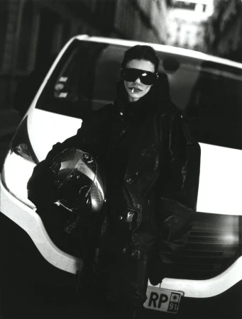Monochrome shot of a stylish adult leaning on a car, cigarette in mouth, sporting shades and a motorcycle helmet, urban setting.