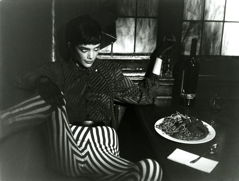 Monochrome portrait of a woman in striped clothing seated at a table with wine and spaghetti, creating an editorial fashion image.