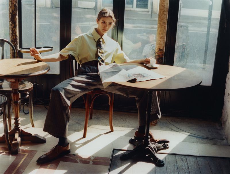 A woman sits at a table in an indoor setting, wearing a collared shirt and tie, with a newspaper, coffee cup, and natural lighting.