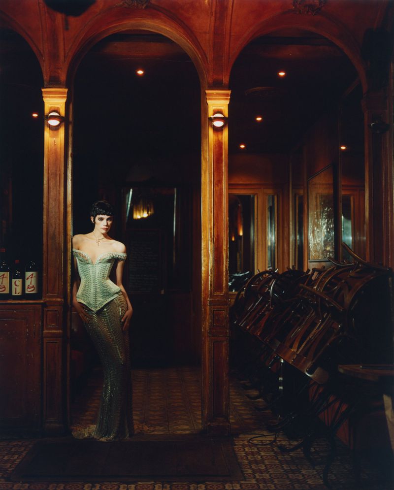 Elegant woman in off-shoulder dress stands in archway of a vintage bar with dark wood paneling and tiled floors under warm lighting.