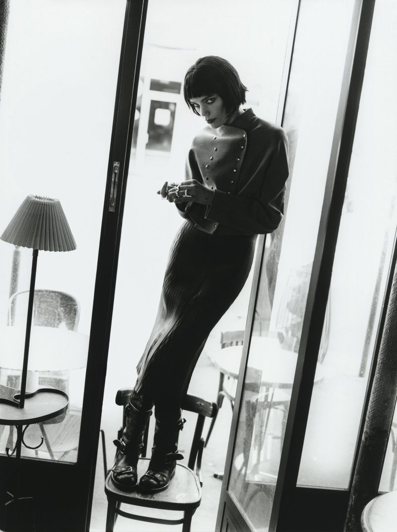Black and white fashion photo of a stylish woman in a jacket and skirt leaning against a window with boots, standing on a chair indoors.