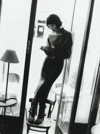 Black and white fashion photo of a stylish woman in a jacket and skirt leaning against a window with boots, standing on a chair indoors.