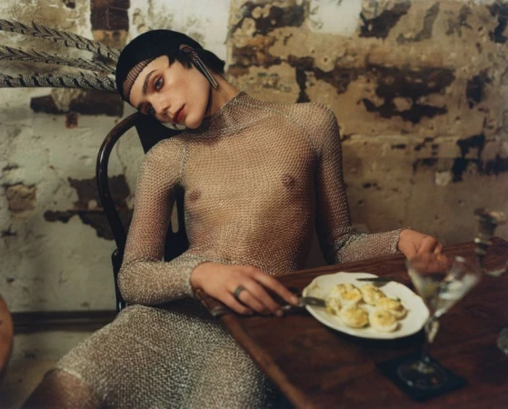 A pale-skinned woman in a chainmail dress sits at a wooden table with a plate of deviled eggs and a martini in a dimly lit, textured room.