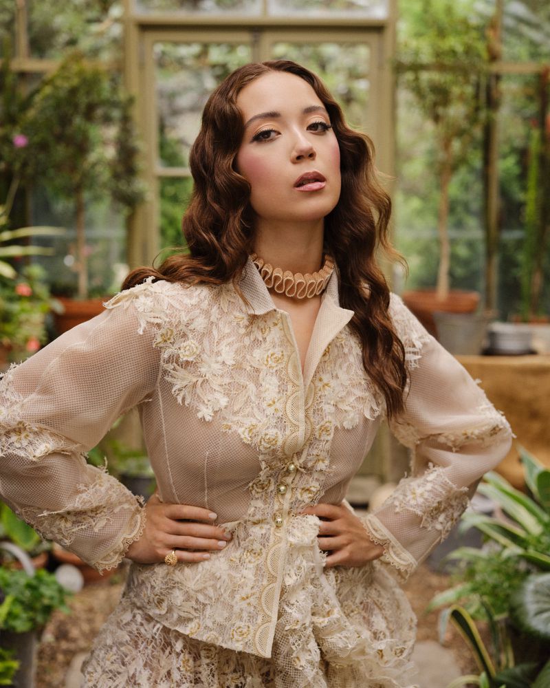 Portrait of a woman in a greenhouse, wearing an ornate beige lace jacket and skirt, with a ruched choker necklace and hands on hips.